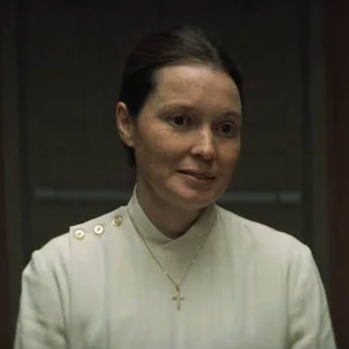 A woman in a formal white outfit with a cross necklace stands indoors, looking attentive