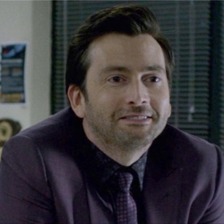 A person in a suit and tie is smiling while leaning on a desk in an office setting with blinds in the background
