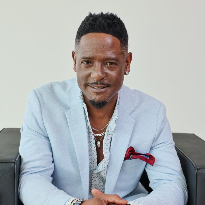 Person seated on a chair wearing a light suit, patterned shirt, and layered necklaces. Hands are clasped, exuding confidence and style