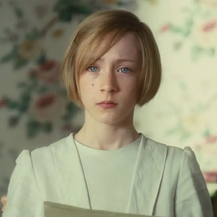 Young person with short hair, wearing a white outfit, stands indoors holding paper, looking contemplative against a floral wallpaper backdrop