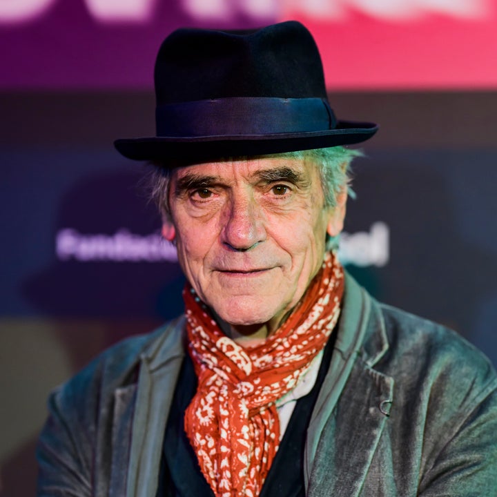I don’t know who this is, but an older man in a hat and scarf wears a textured blazer and vest, standing on a red carpet