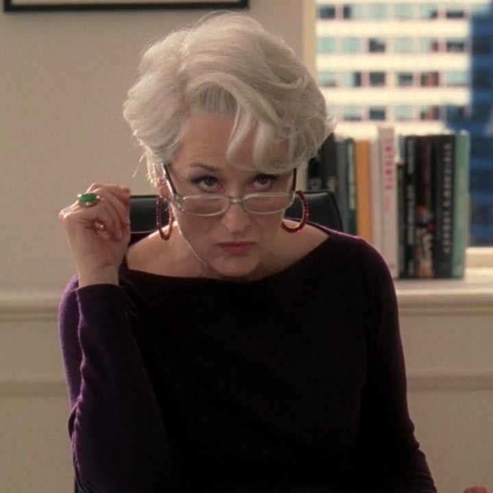 A person with short, white hair sits at a desk, looking over glasses with a serious expression. They hold a sheet of paper and wear a dark top