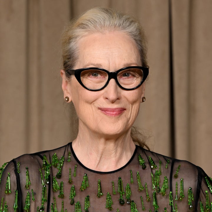 Person wearing black glasses and a sheer dress with green embellishments smiles subtly against a neutral backdrop
