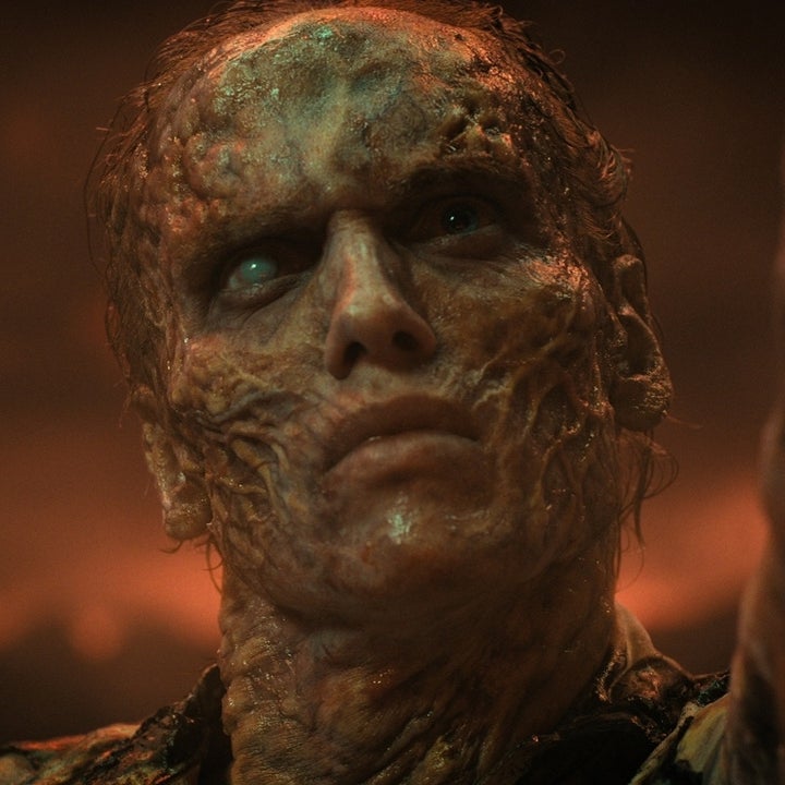 A person with a heavily scarred and burned face looks upward with a serious expression, set against a smoky backdrop