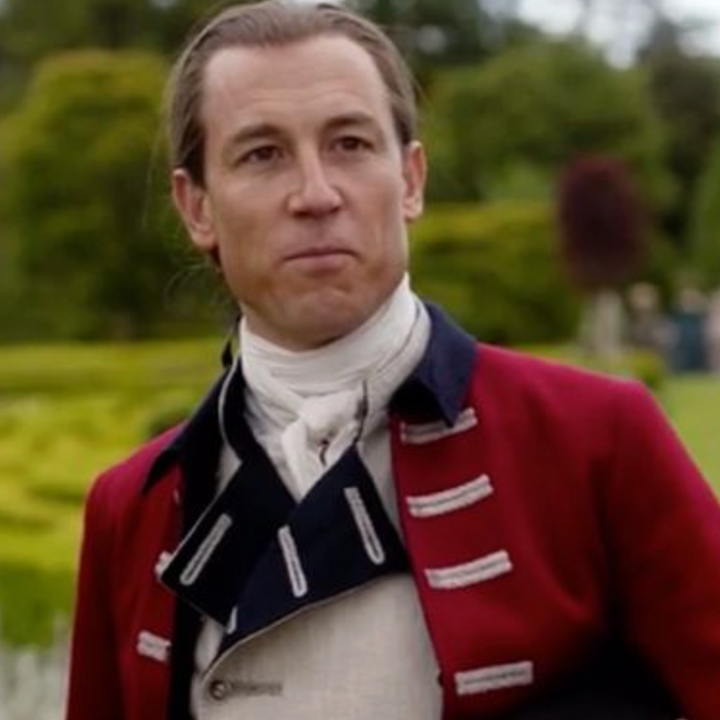 A person in a historical military-style costume with a white cravat and red coat stands in a garden setting