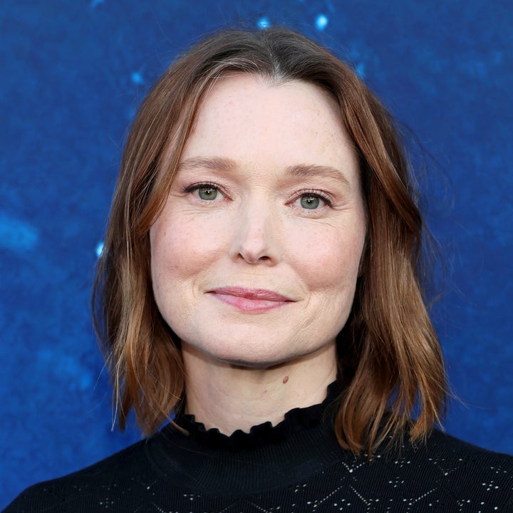 A person with shoulder-length hair wears a black top with a patterned texture, standing against a blue backdrop