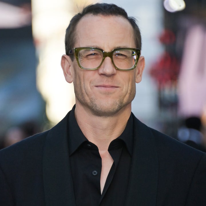 A person in a dark suit jacket and glasses stands outdoors, smiling slightly