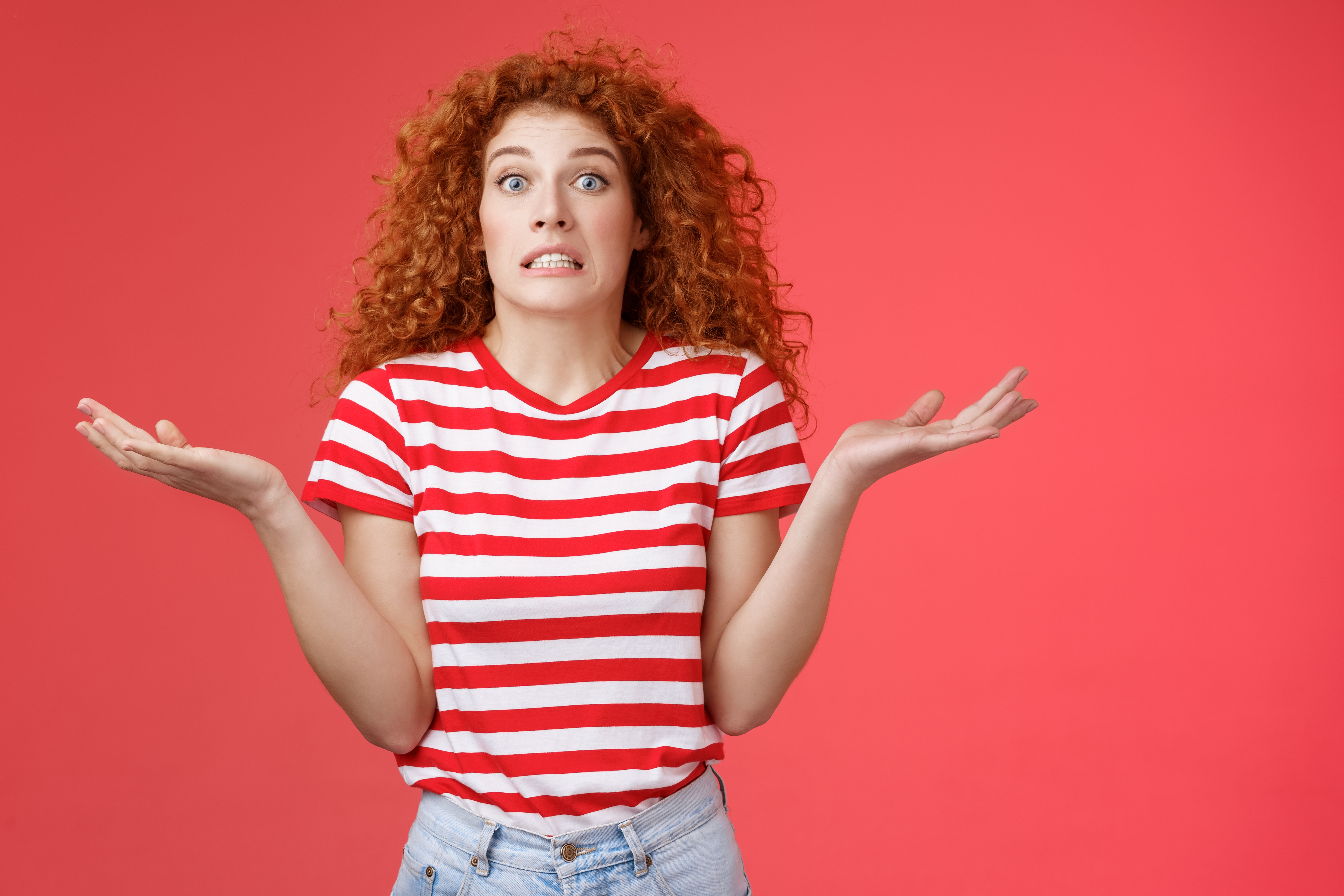 Person with curly hairsbreadth  shrugs with a amazed  expression, wearing a striped shirt, against a plain background