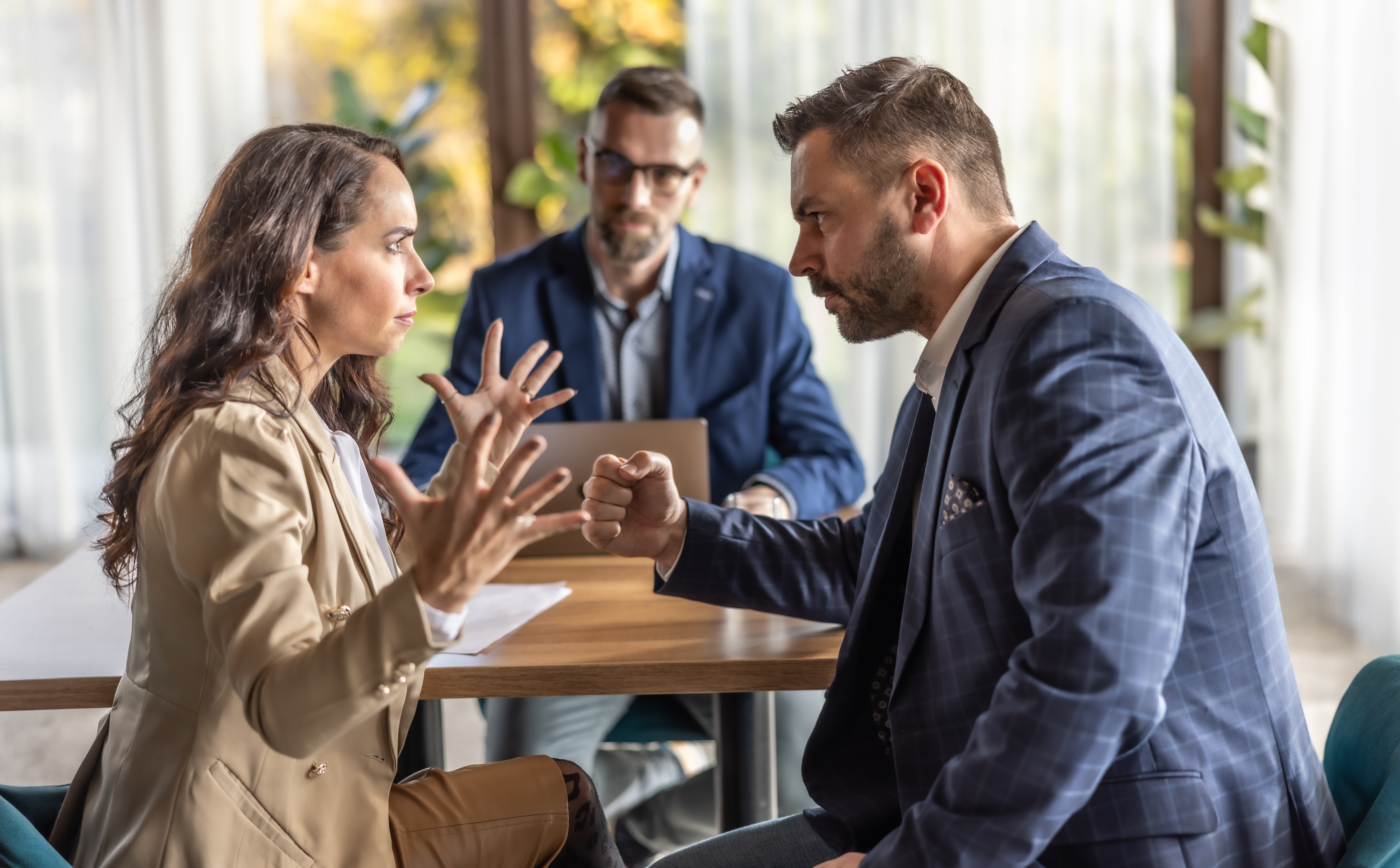 Two radical   successful  a heated treatment  astatine  a table, facing each   different   with aggravated  expressions. A 3rd  idiosyncratic   observes them, holding a laptop