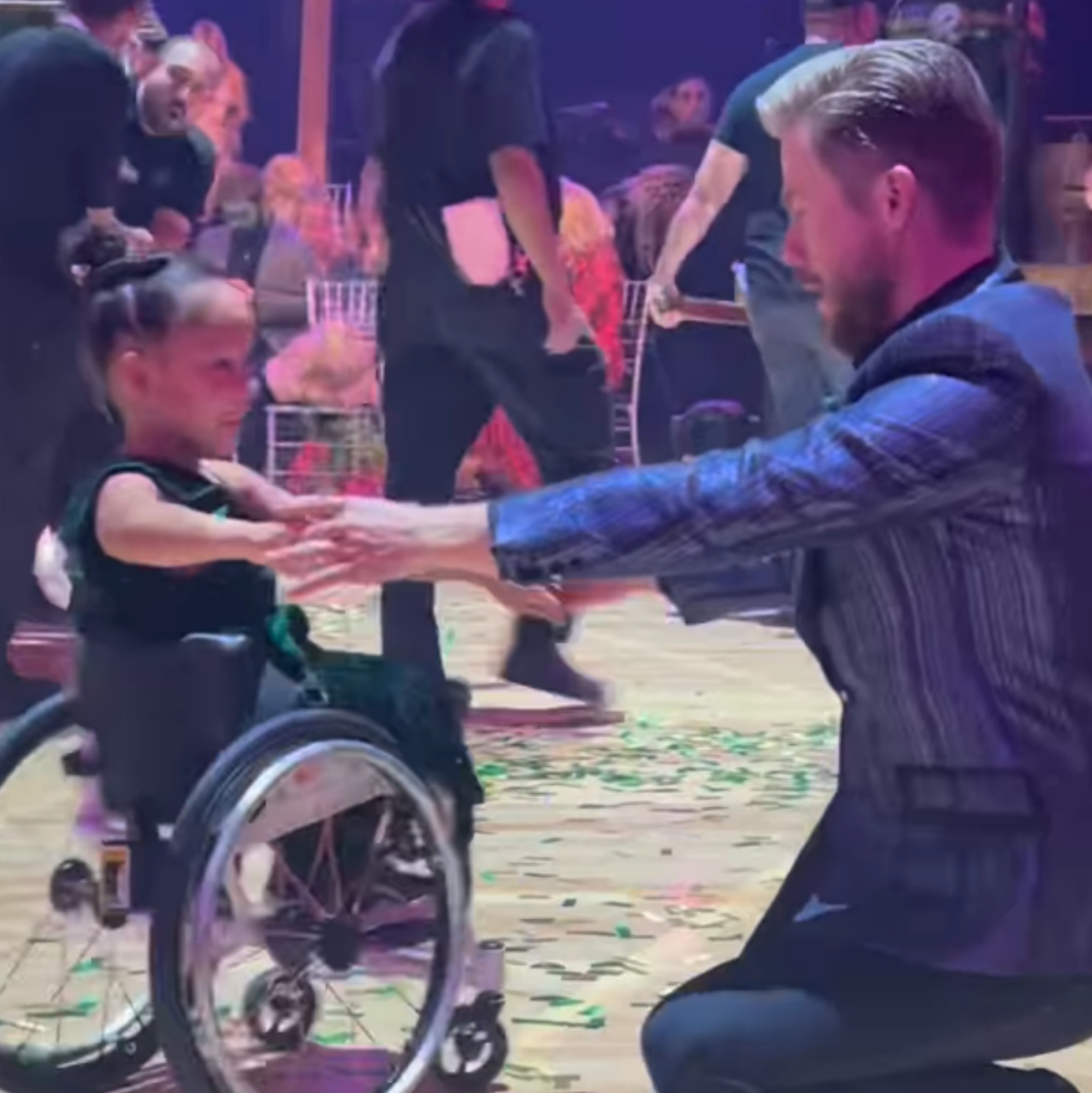 Man kneels and extends arms to a smiling young girl in a wheelchair on a crowded dance floor