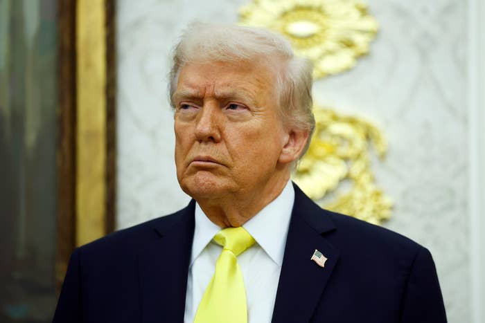 A person in a suit with an American flag pin stands indoors, looking to the side with a serious expression