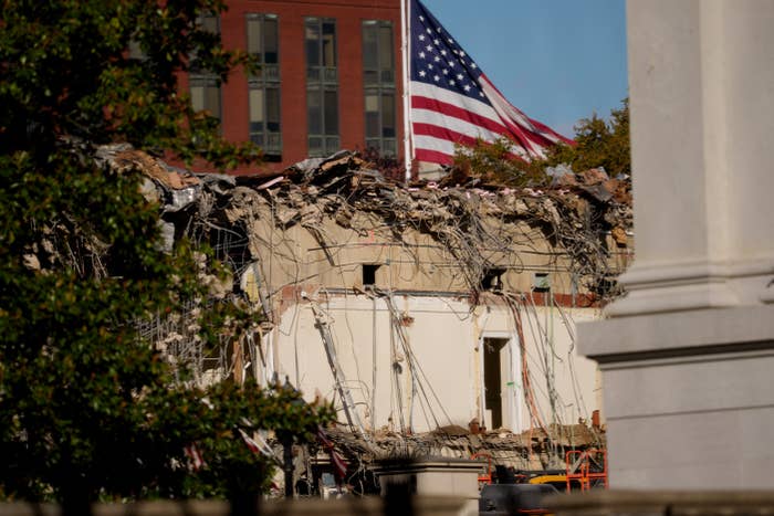 A partially demolished building with exposed wires and debris stands in front of a waving American flag and a brick building in the background