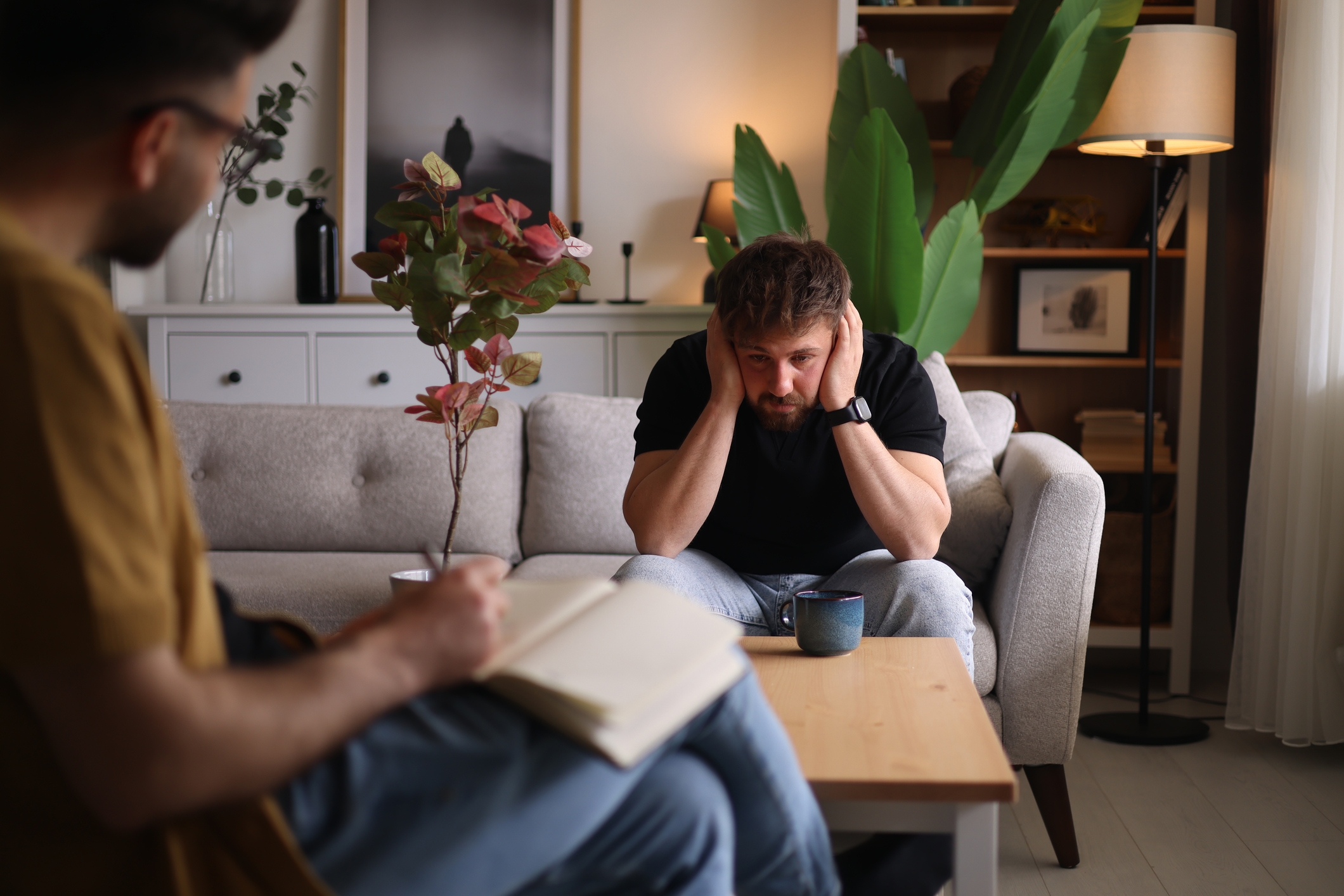 To personer sitter i en stue. En person er synlig bekymret, med hodet i hendene, mens den andre tar notater og foreslår en terapiøkt