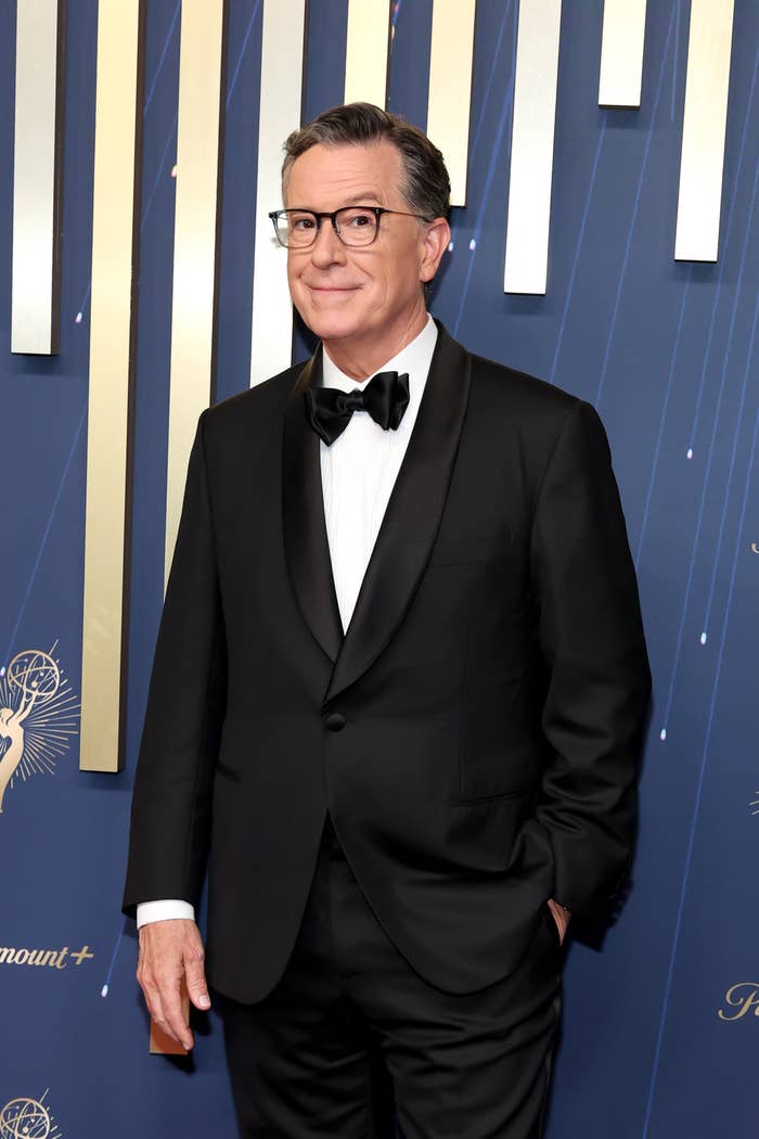 A person in a formal black tuxedo on a red carpet, posing confidently with one hand in their pocket against a patterned backdrop