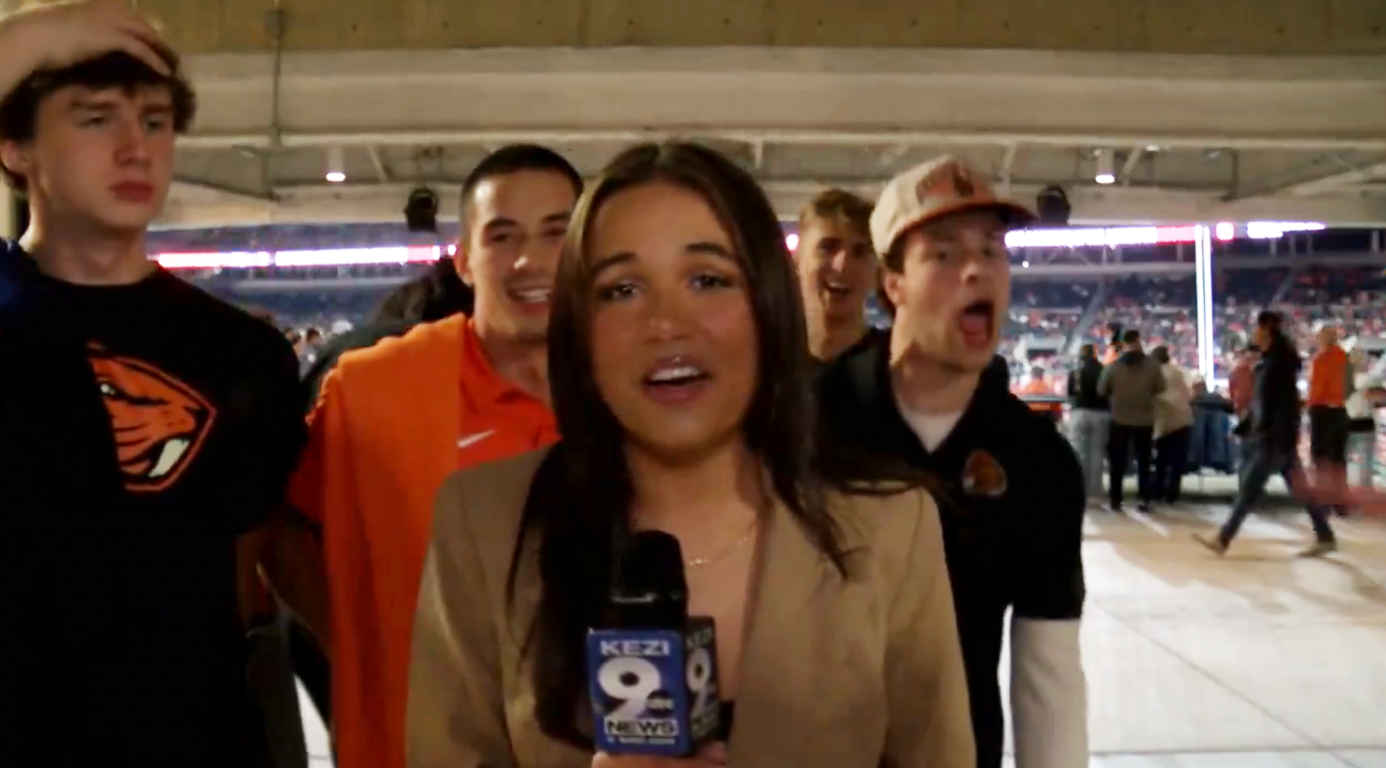 Reporter holding a microphone on a sports field with excited fans making faces in the background