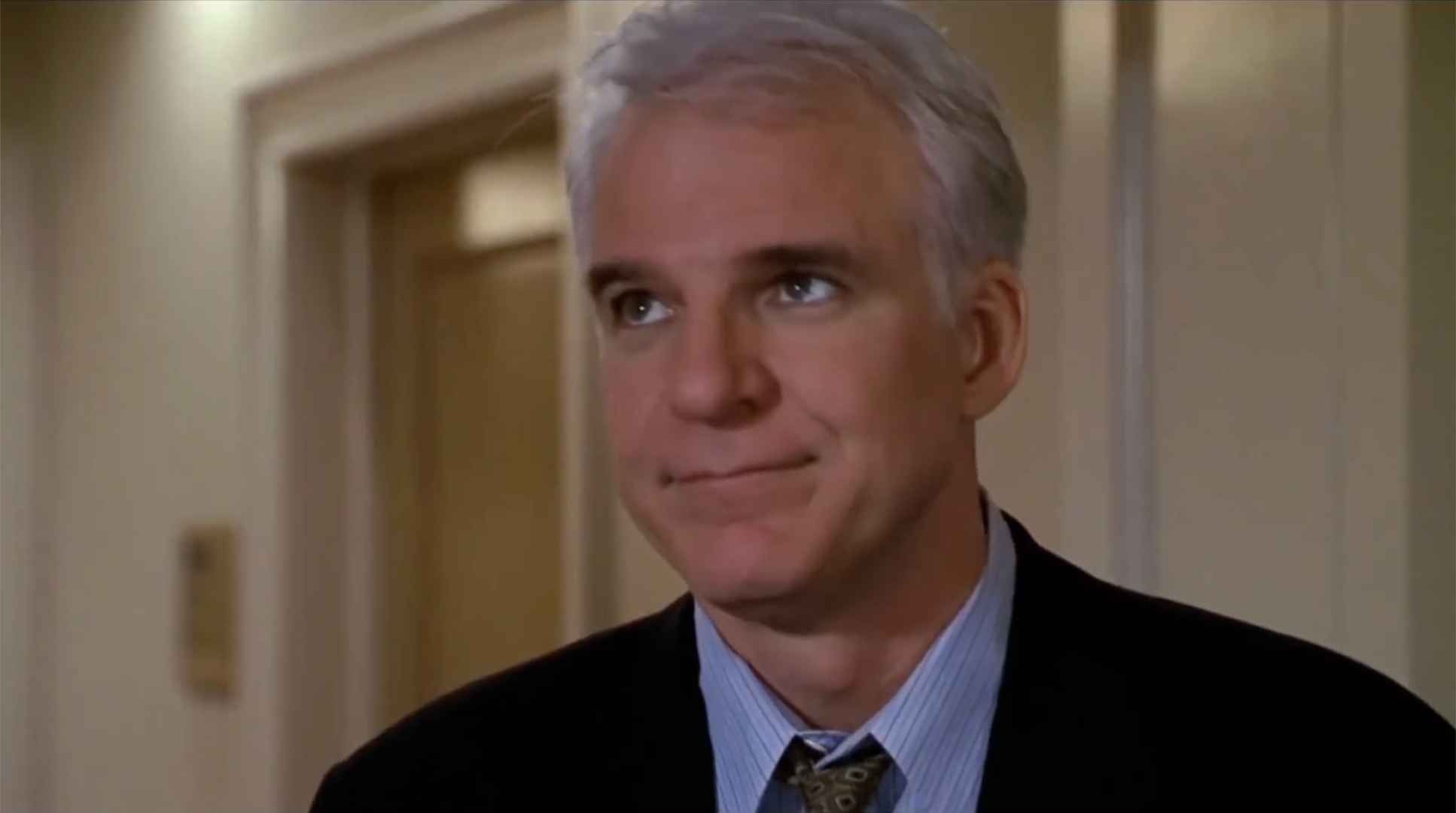 Man successful  a suit   with a striped garment  and patterned necktie  stands indoors, looking somewhat  upward with a contemplative expression