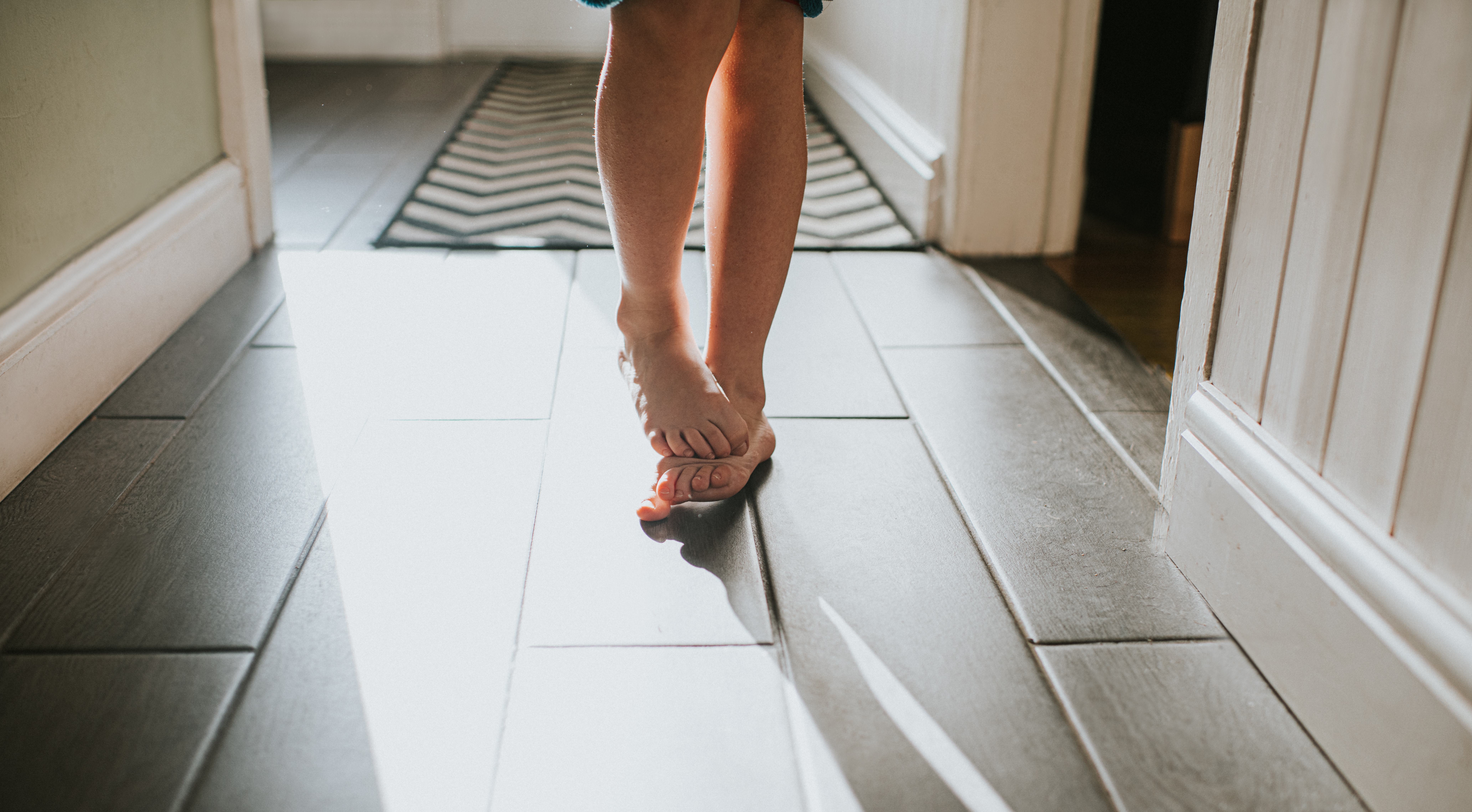 En person som går barbeint nedover en solbelyst gang, og føttene kaster skygger på et tregulv