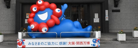 建物の前に笑顔の青いマスコットが展示され、看板には「みなさまのご協力に感謝!大阪・関西万博」。