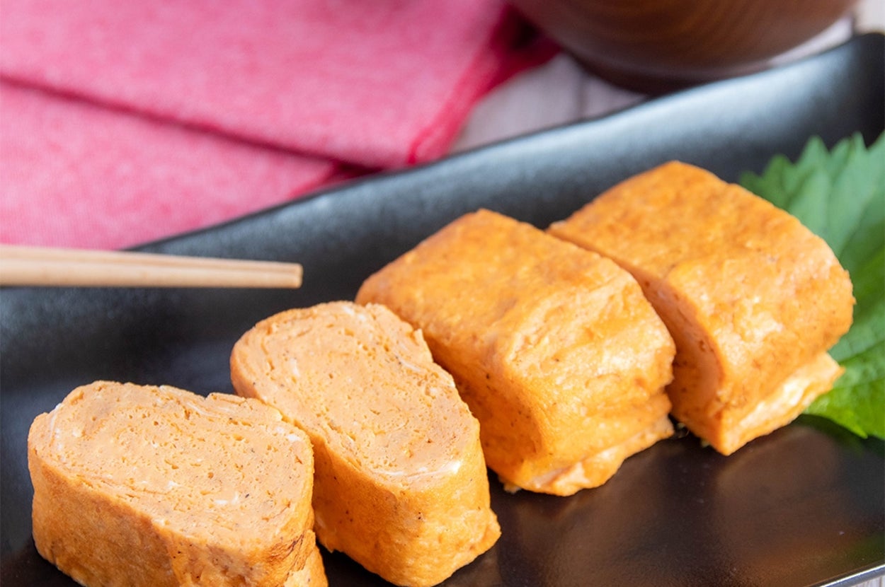 黒い皿に盛られた4切れの卵焼き。左側に箸、右上に味噌汁。お皿に葉が添えられている。