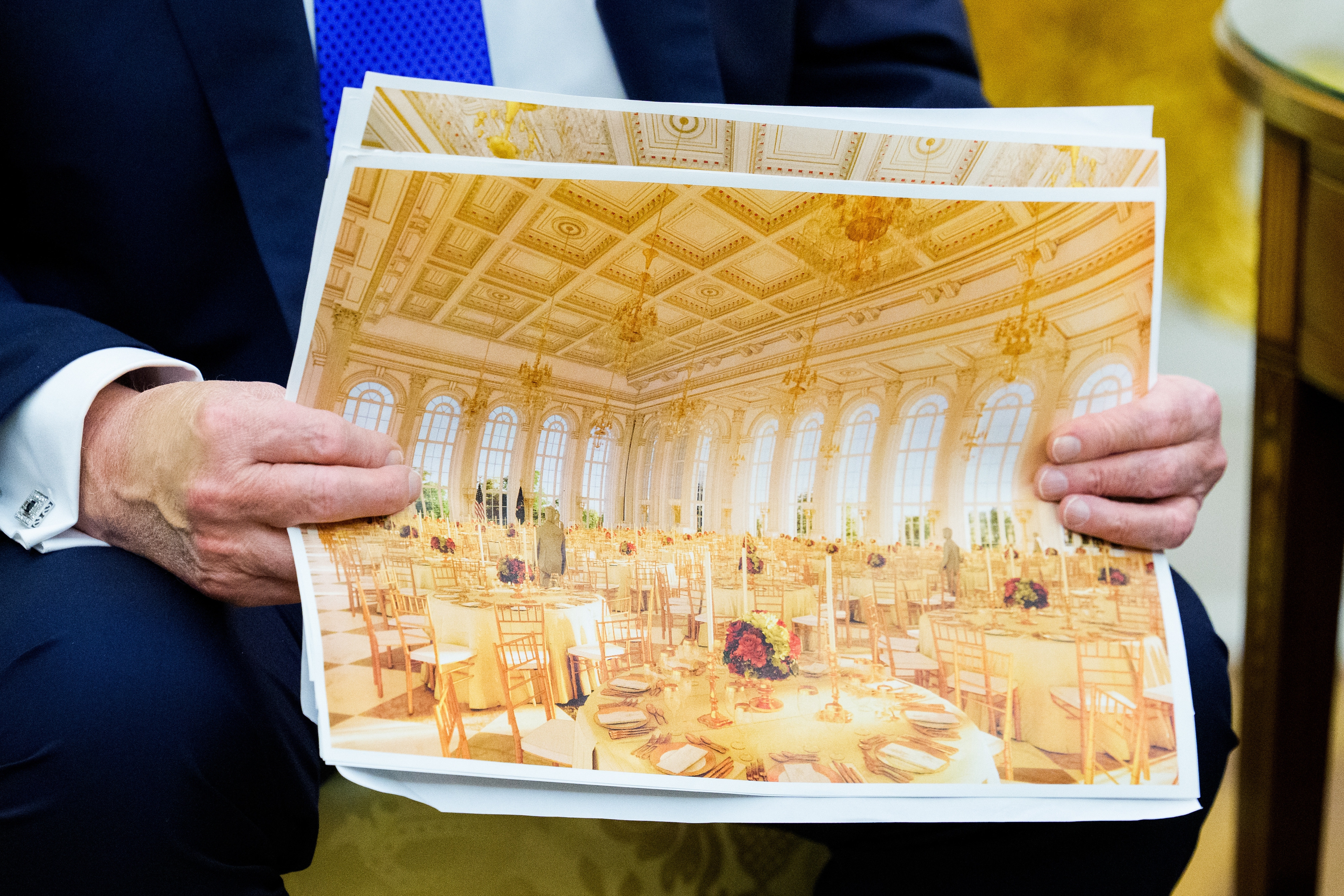 Person holding a photo of a luxurious, ornate ballroom set for an event with elegant table settings and chandeliers