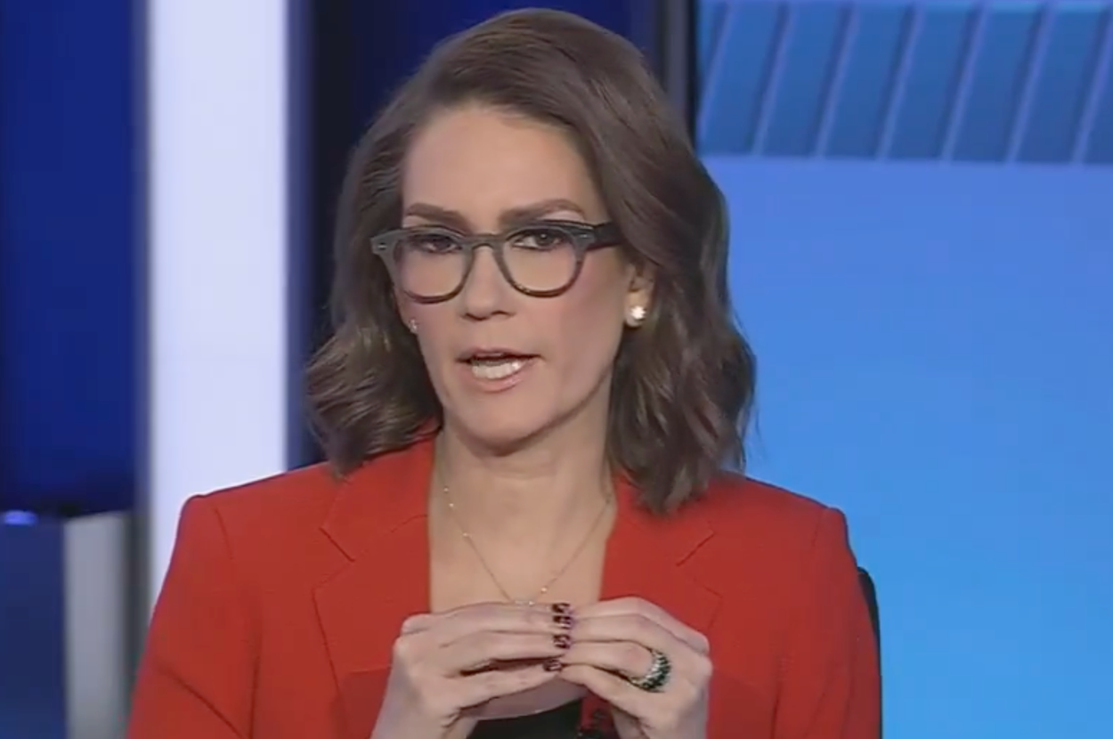 A person with glasses in a studio setting, wearing a tailored suit jacket, appears to be speaking on camera