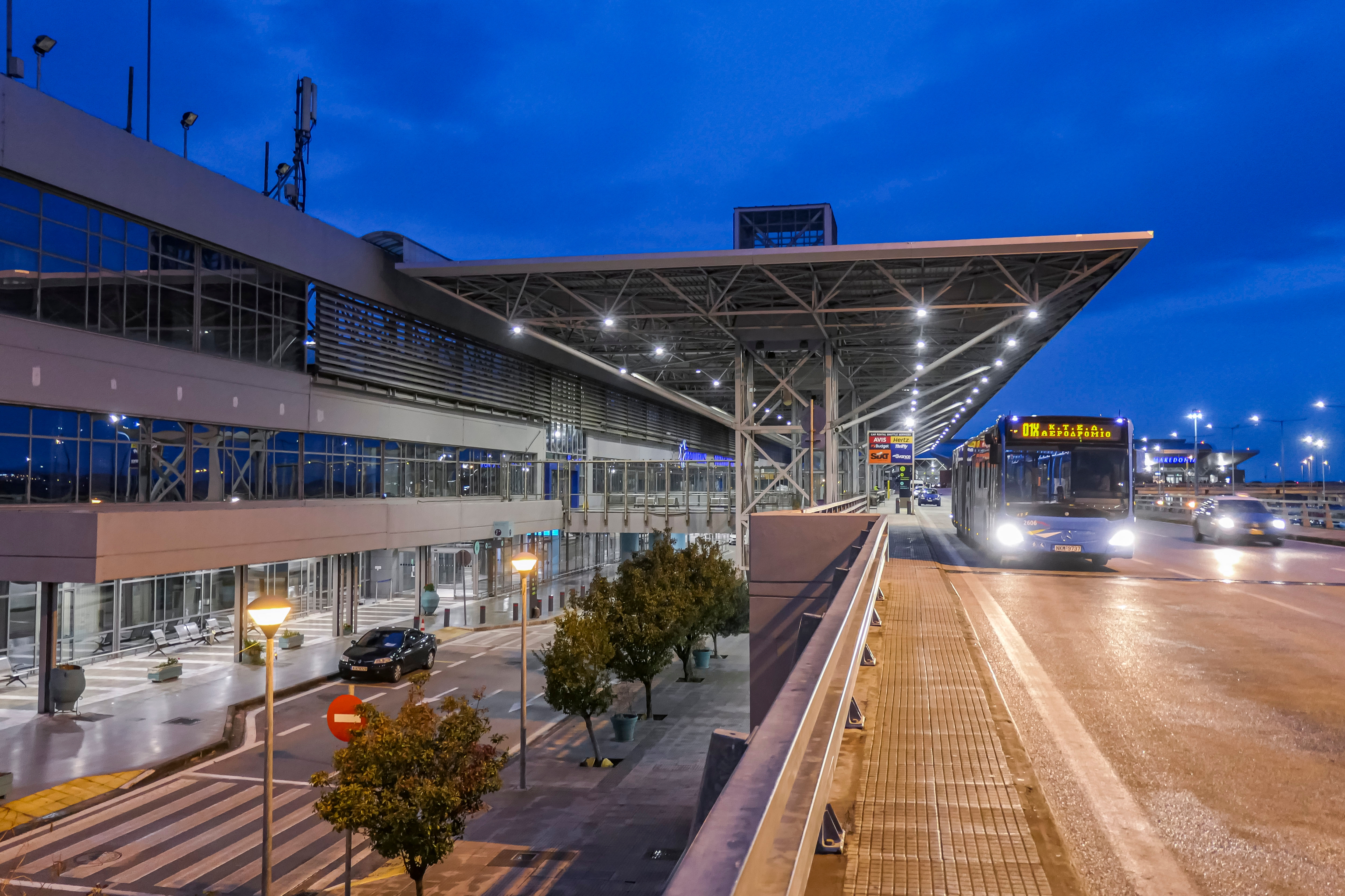 En moderne flyplassterminal om natten med en godt opplyst buss som ankommer, trær langs veien og en klar himmel over