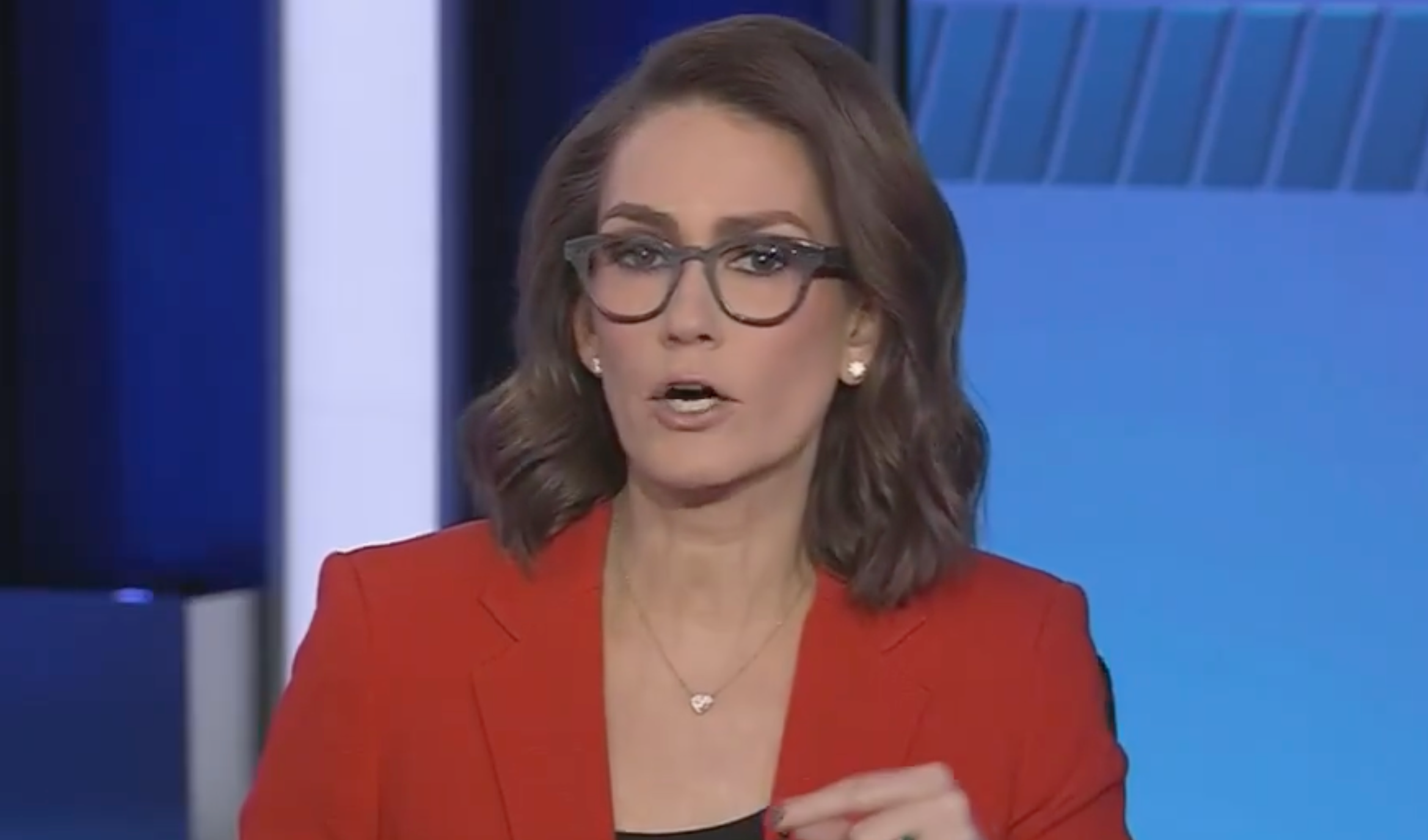 A person wearing glasses and a red blazer speaks in a news studio, appearing engaged in a discussion