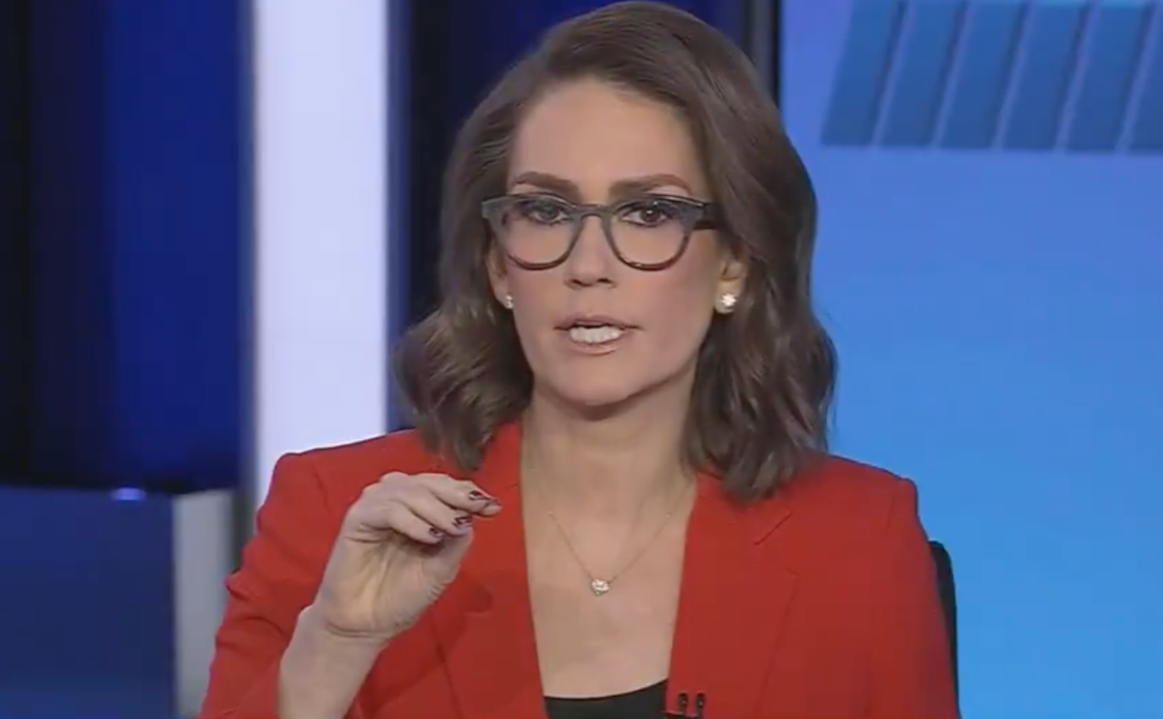 News anchor in a studio setting, wearing glasses and a red blazer, gesturing while speaking on a broadcast