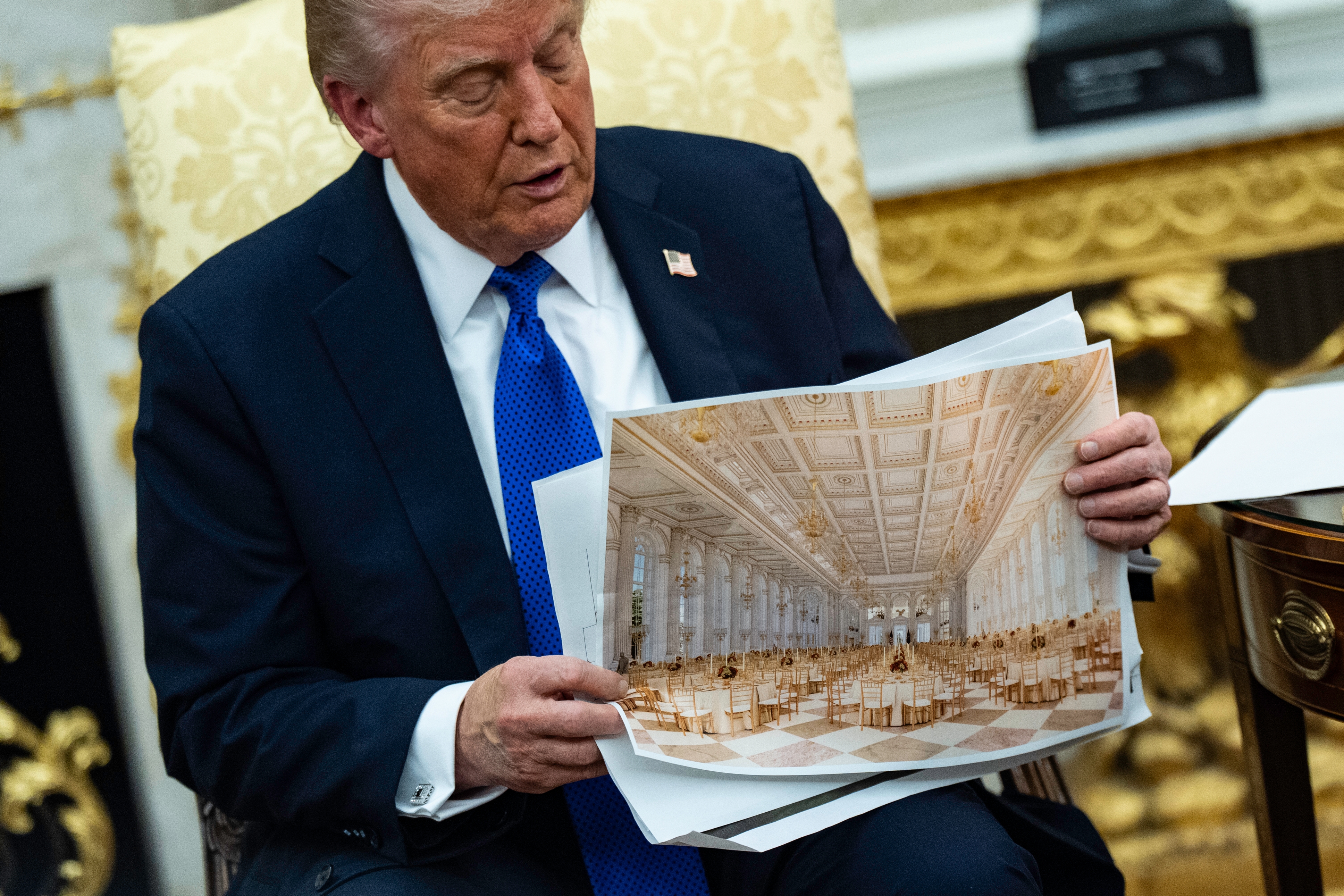 A person in a suit holding a photo of a grand, empty banquet hall, seated in an ornate room