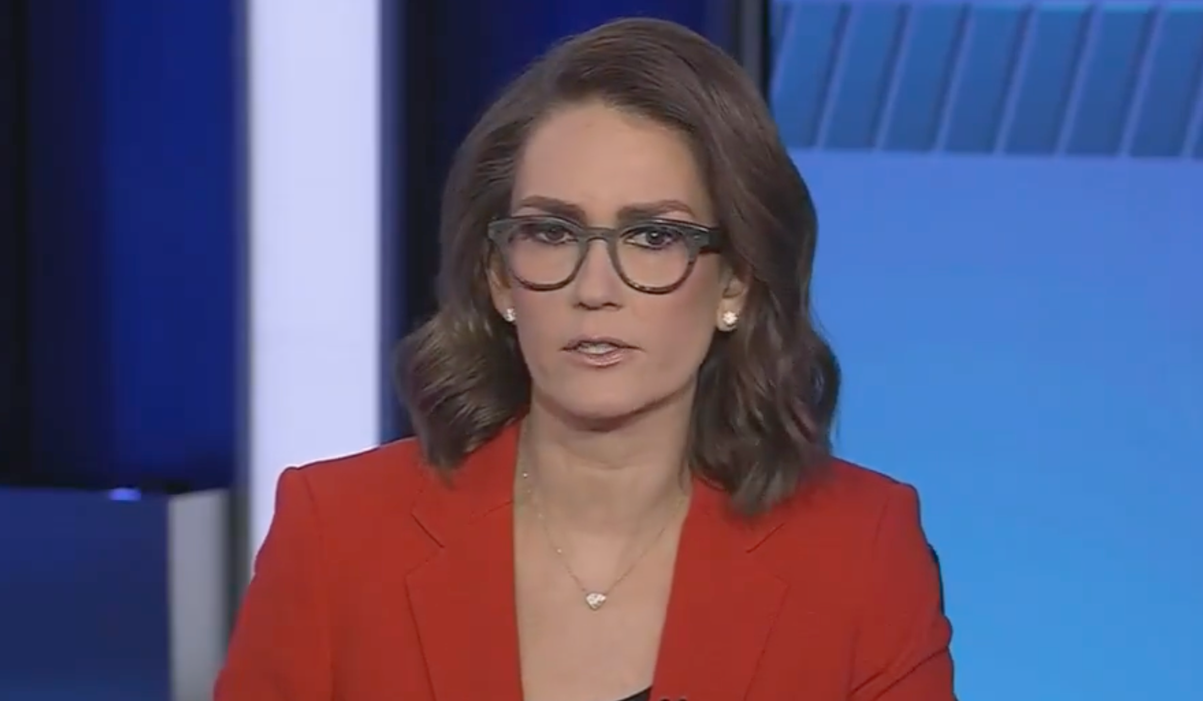 Person wearing glasses and a blazer appears on a news broadcast set, looking directly at the camera