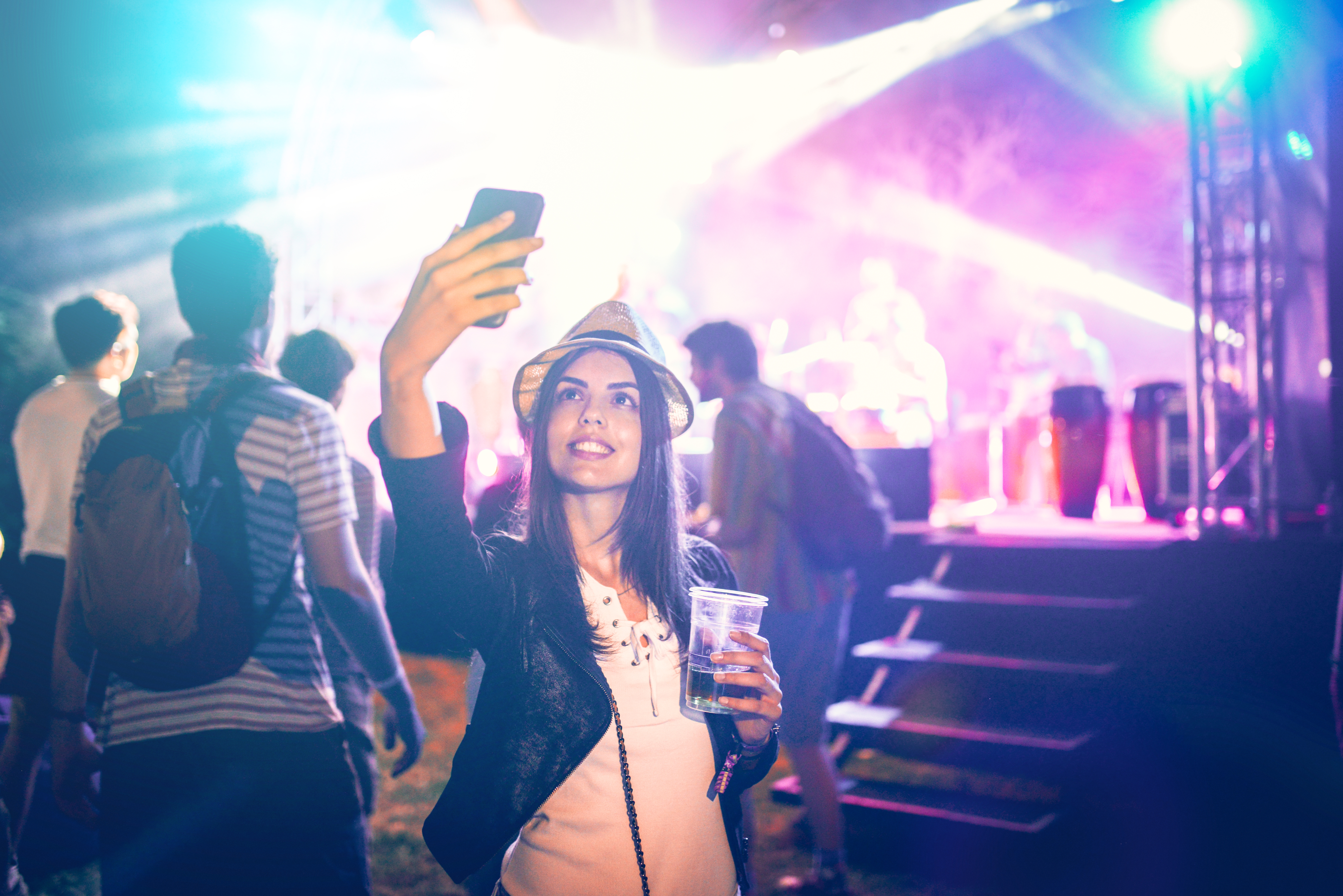 En person i en stilig hatt tar en selfie på en livlig utendørskonsert, med en drink i hånden og skarpe scenelys i bakgrunnen