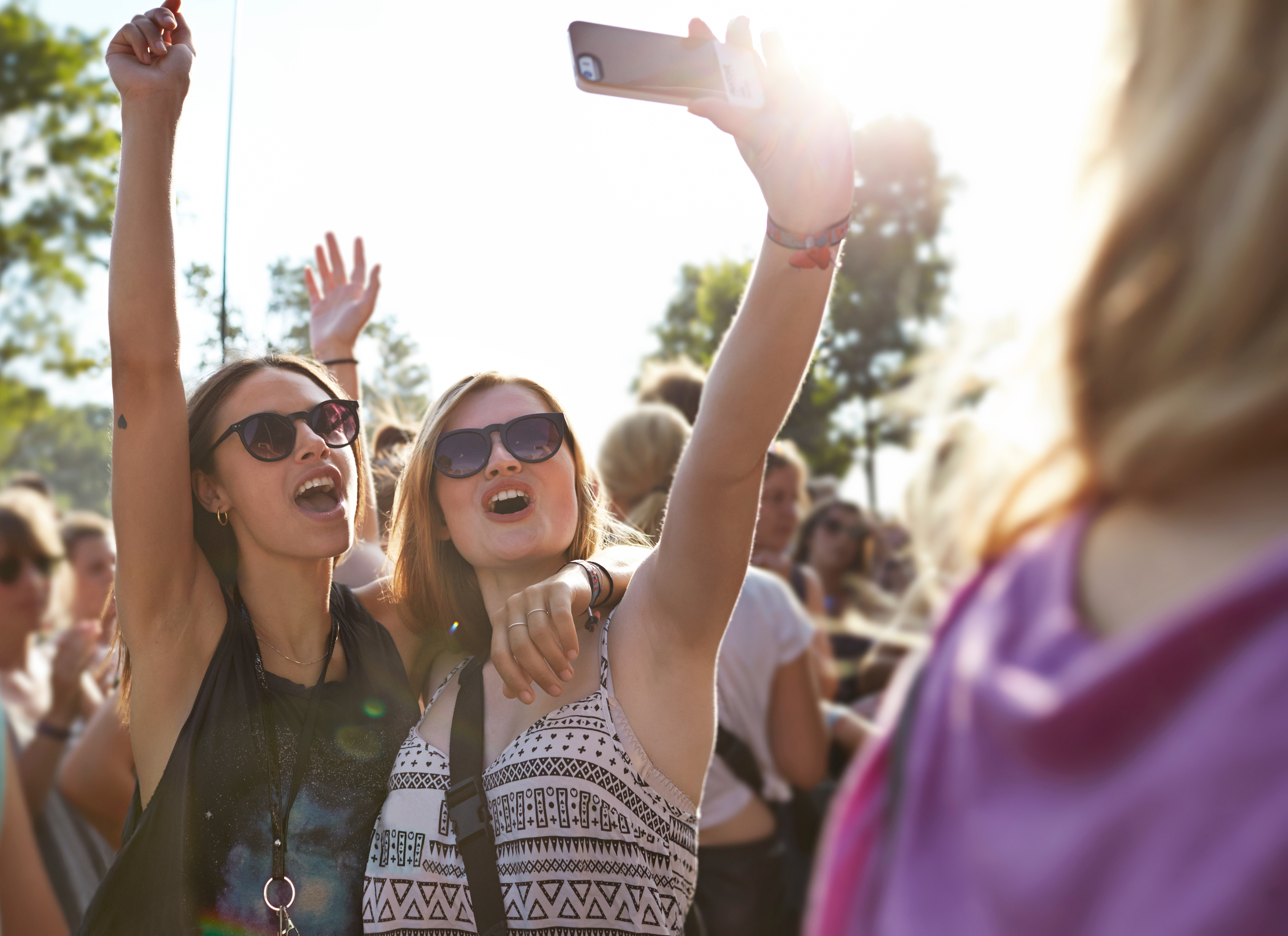 To personer i solbriller tar en selfie på et utendørsarrangement, med en folkemengde i bakgrunnen som nyter solskinnsdagen