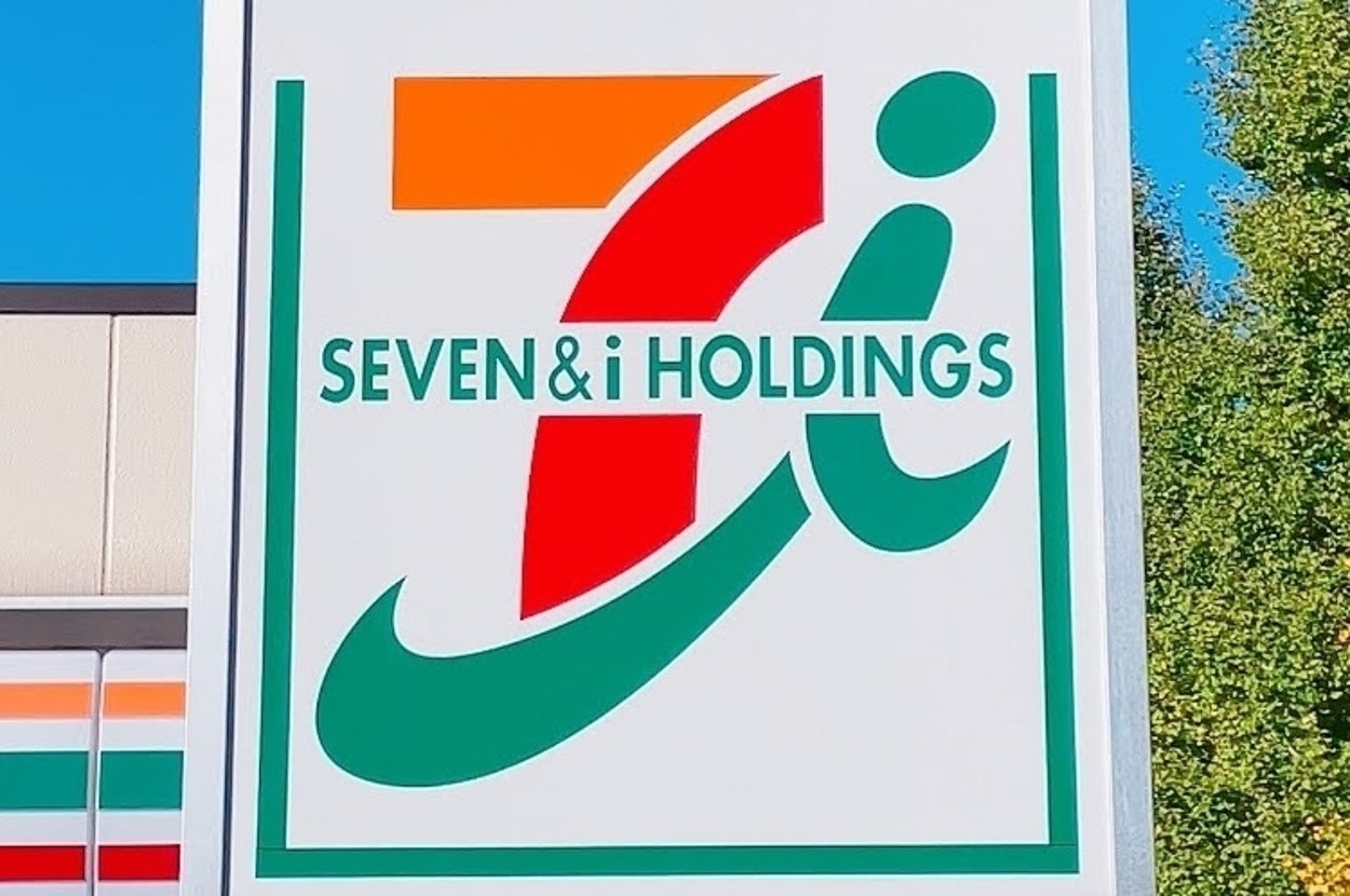 セブン-イレブンの看板。ブランドロゴに「SEVEN & i HOLDINGS」と書かれている。背景に木々が見える。