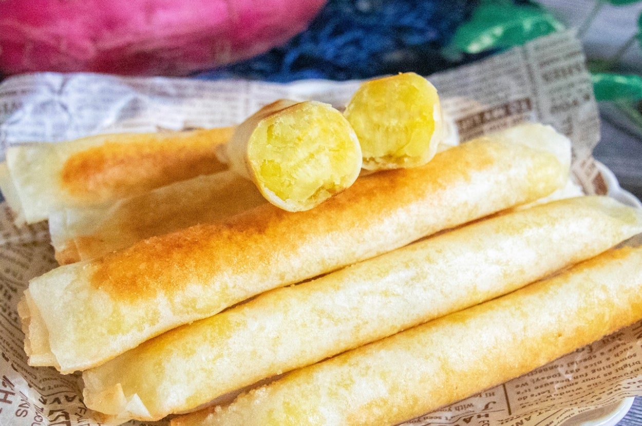 お皿に盛られた細長いスイートポテト春巻き。後ろにさつまいもが置かれている。