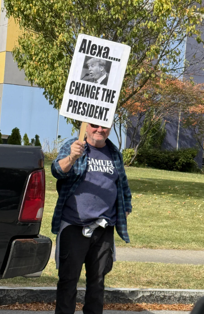 Person holding a sign with a message requesting Alexa to "Change the President," standing by a vehicle