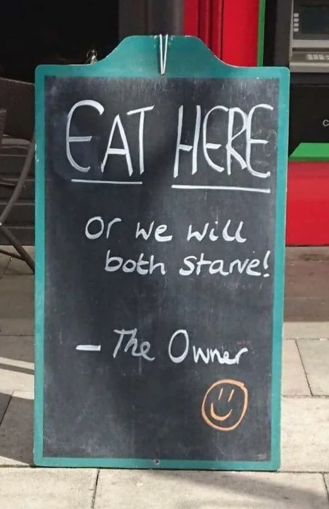 Chalkboard sign reads: "EAT HERE or we will both starve! - The Owner" with a smiley face at the bottom