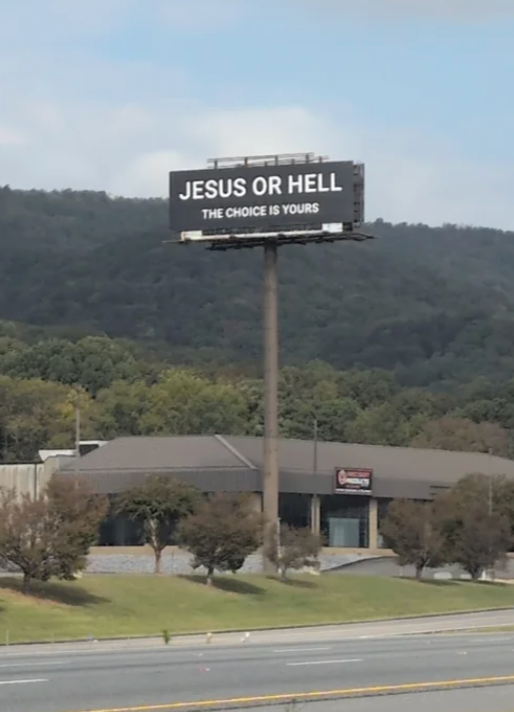 Billboard reads, "JESUS OR HELL. THE CHOICE IS YOURS" against a backdrop of trees and a building