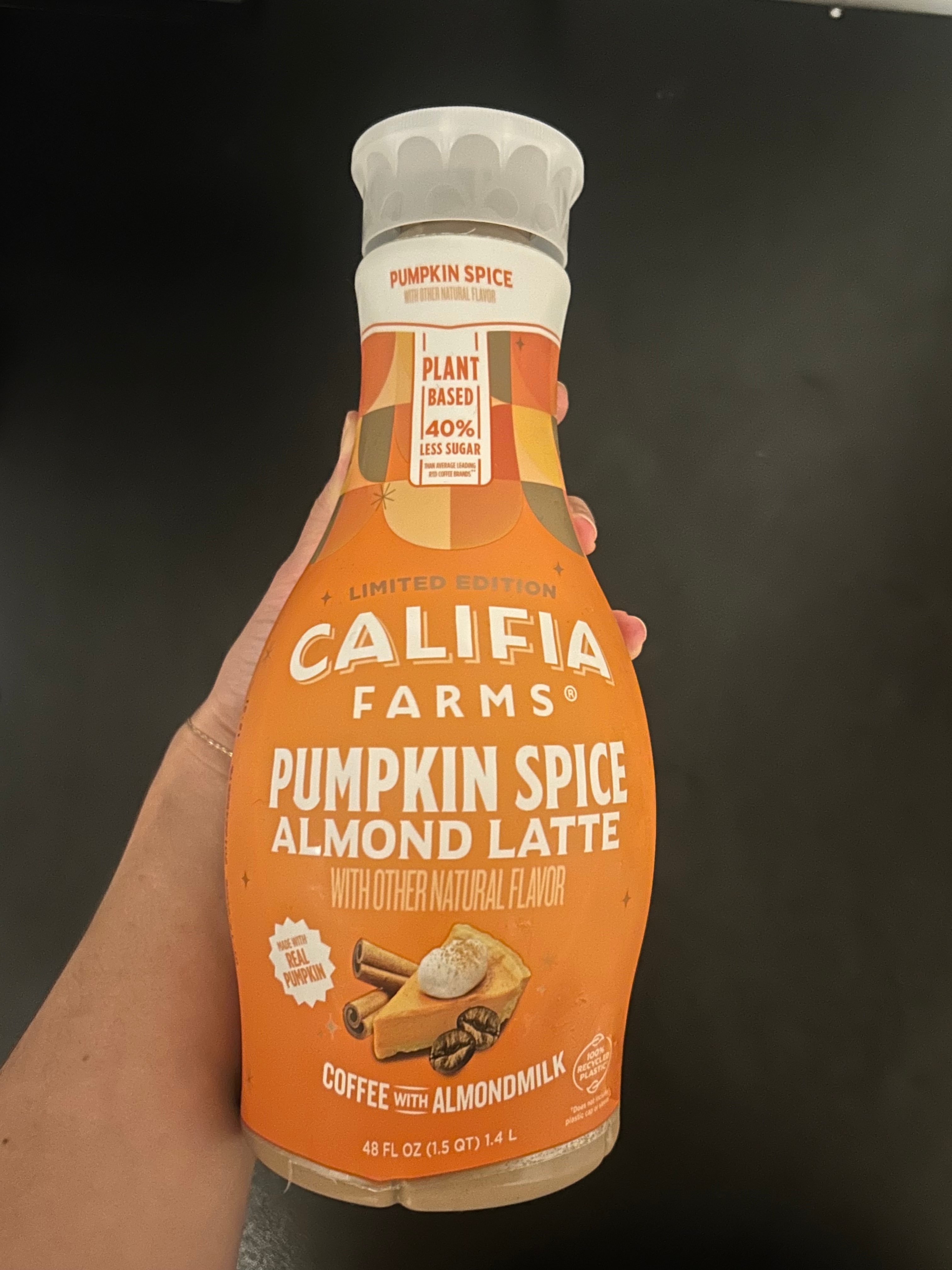 Califia Farms Pumpkin Spice Almond Latte bottle held against a dark background. The label highlights plant-based, limited edition, and less sugar features