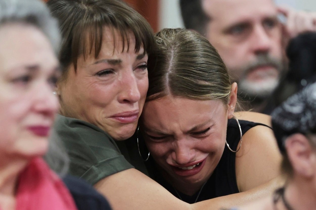 Two women are embracing, visibly emotional and teary-eyed, in a crowded room. Others around them appear somber