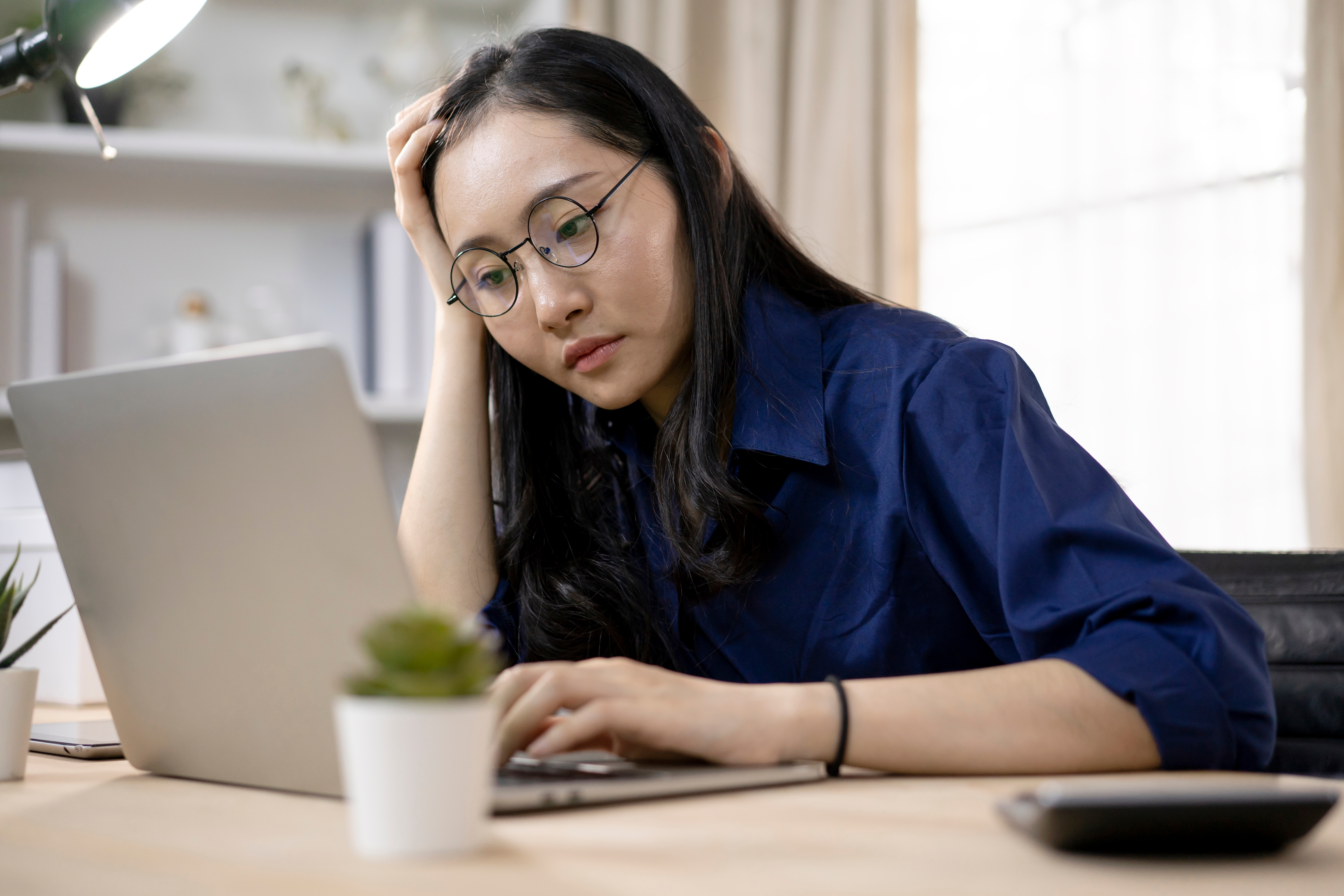 Person med briller, i uformelle klær, fokusert og jobber på en bærbar datamaskin ved et skrivebord med planter