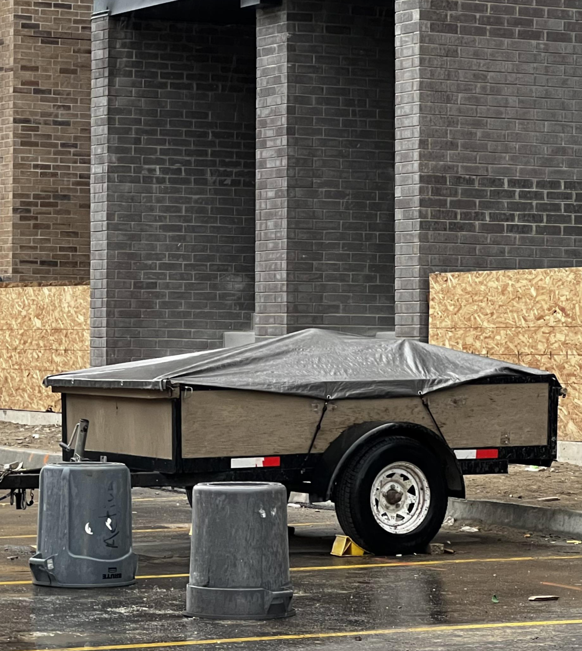 A flatbed trailer covered with a tarp is parked adjacent   a gathering  nether  construction, surrounded by trash cans and scattered debris