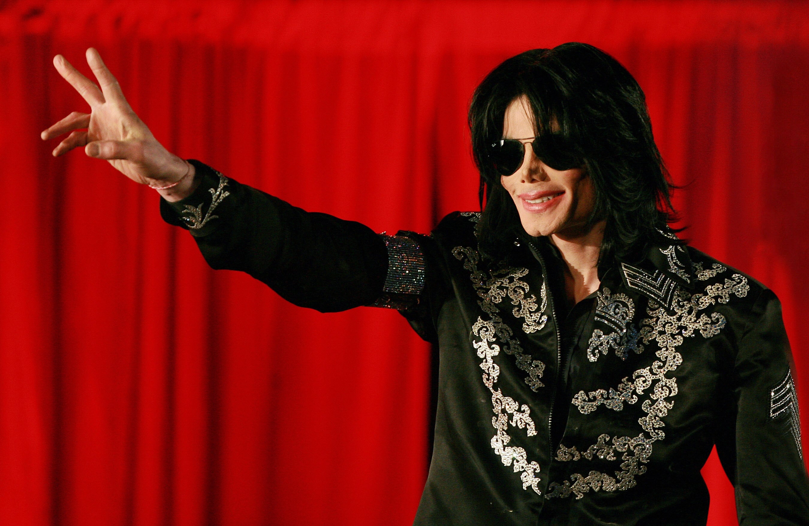 Person in ornate jacket with embroidered details and sunglasses, making a peace sign gesture on a red backdrop