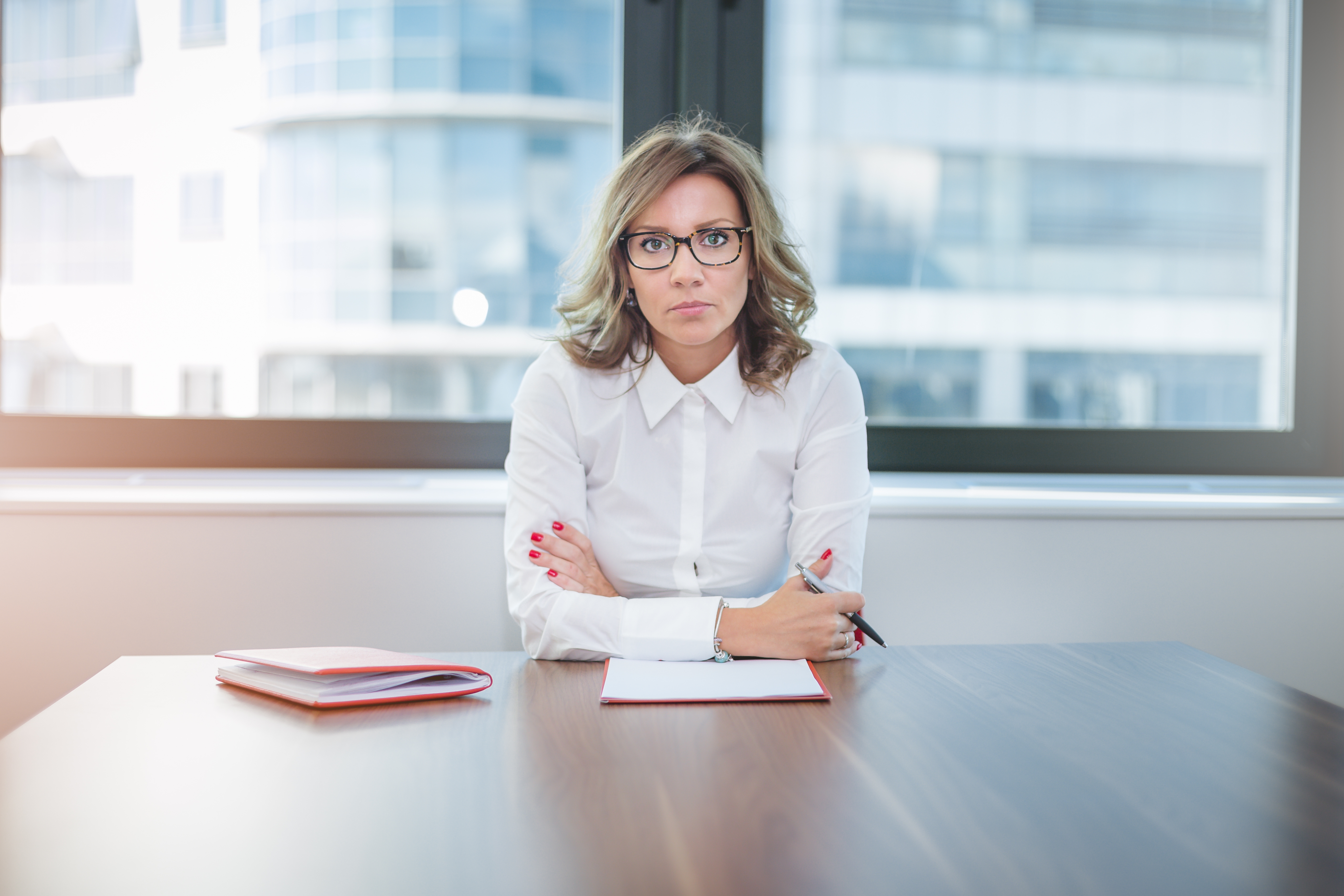 En person i en hvit skjorte sitter ved et bord med foldede armer og ser direkte på kameraet. En notatbok og en penn ligger på bordet