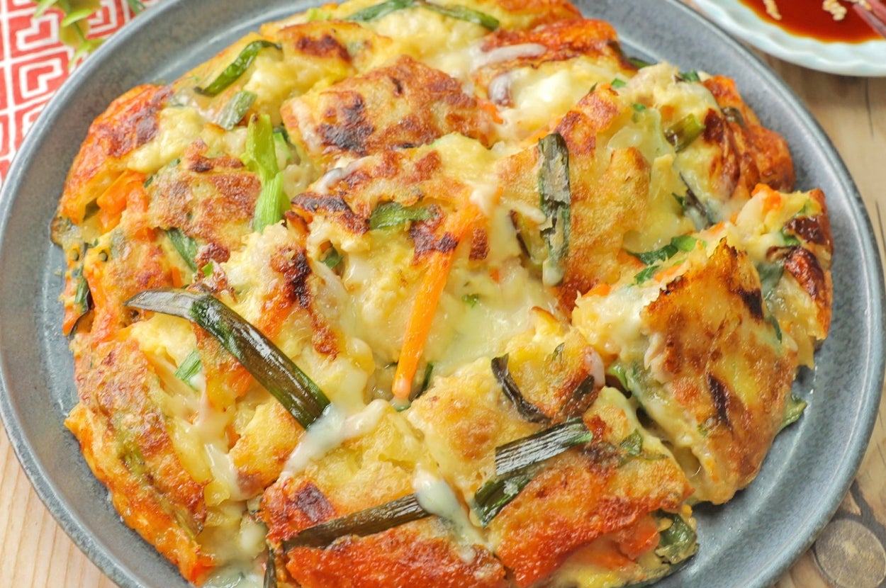 野菜が入ったチヂミが皿に載せられ、隣にタレが入った小皿がある。背景には葉と布が配置されている。