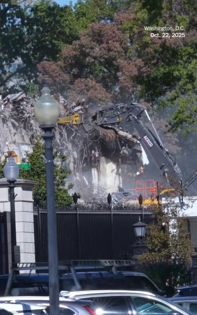 Building demolition in progress in Washington, D.C., with an excavator tearing down the structure. Trees and street lamps visible in the foreground