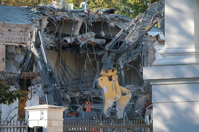 Demolition of a building underway, with machinery breaking down walls. Debris and exposed wires are visible, showing extensive deconstruction progress