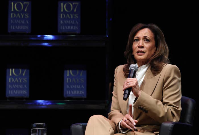 Kamala Harris in a tan suit speaks into a microphone at an event, seated on a stage with books titled &quot;107 Days&quot; on a shelf behind her