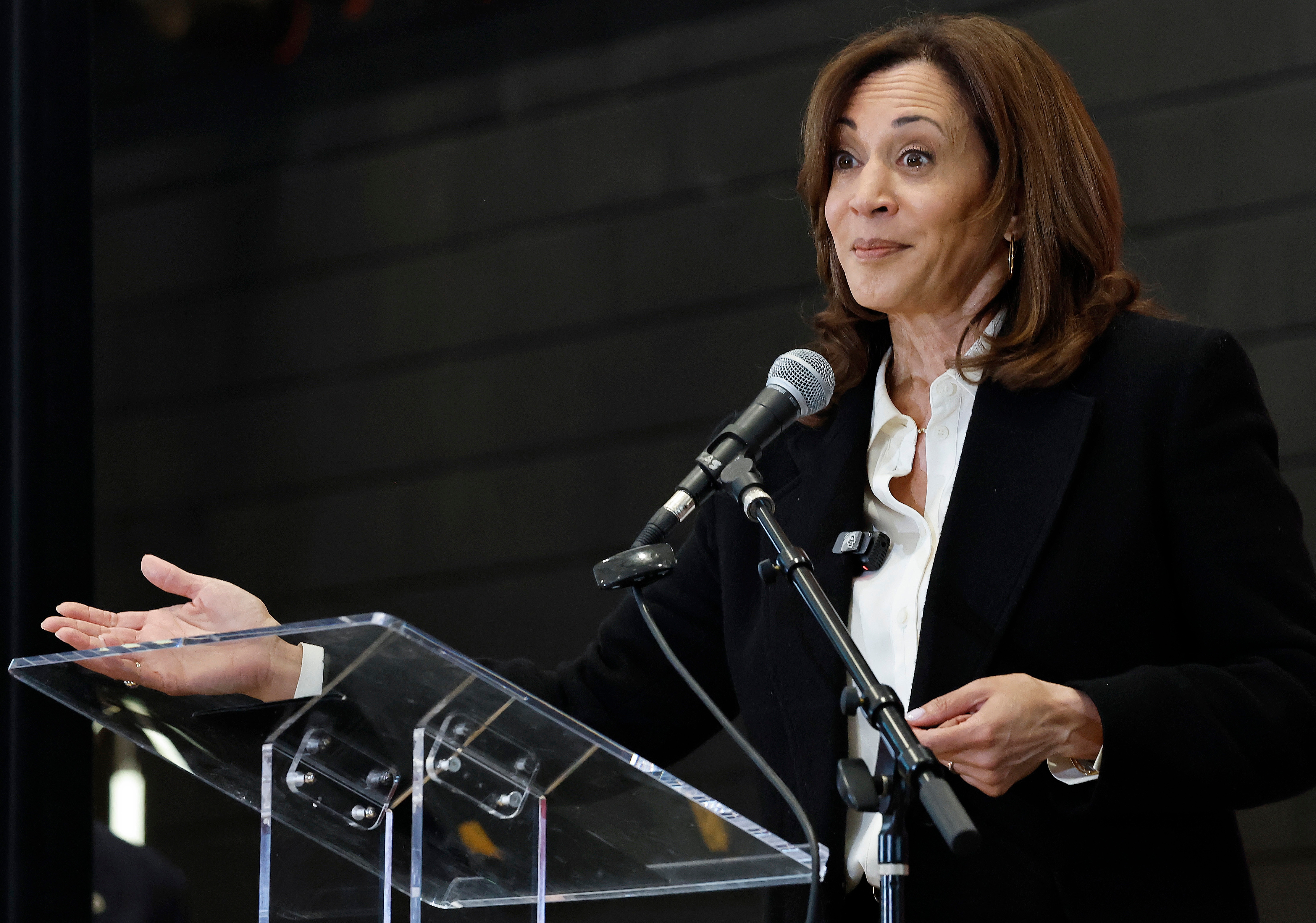 Kamala Harris at a podium with a microphone, gesturing while speaking. They are wearing a formal black jacket over a white shirt