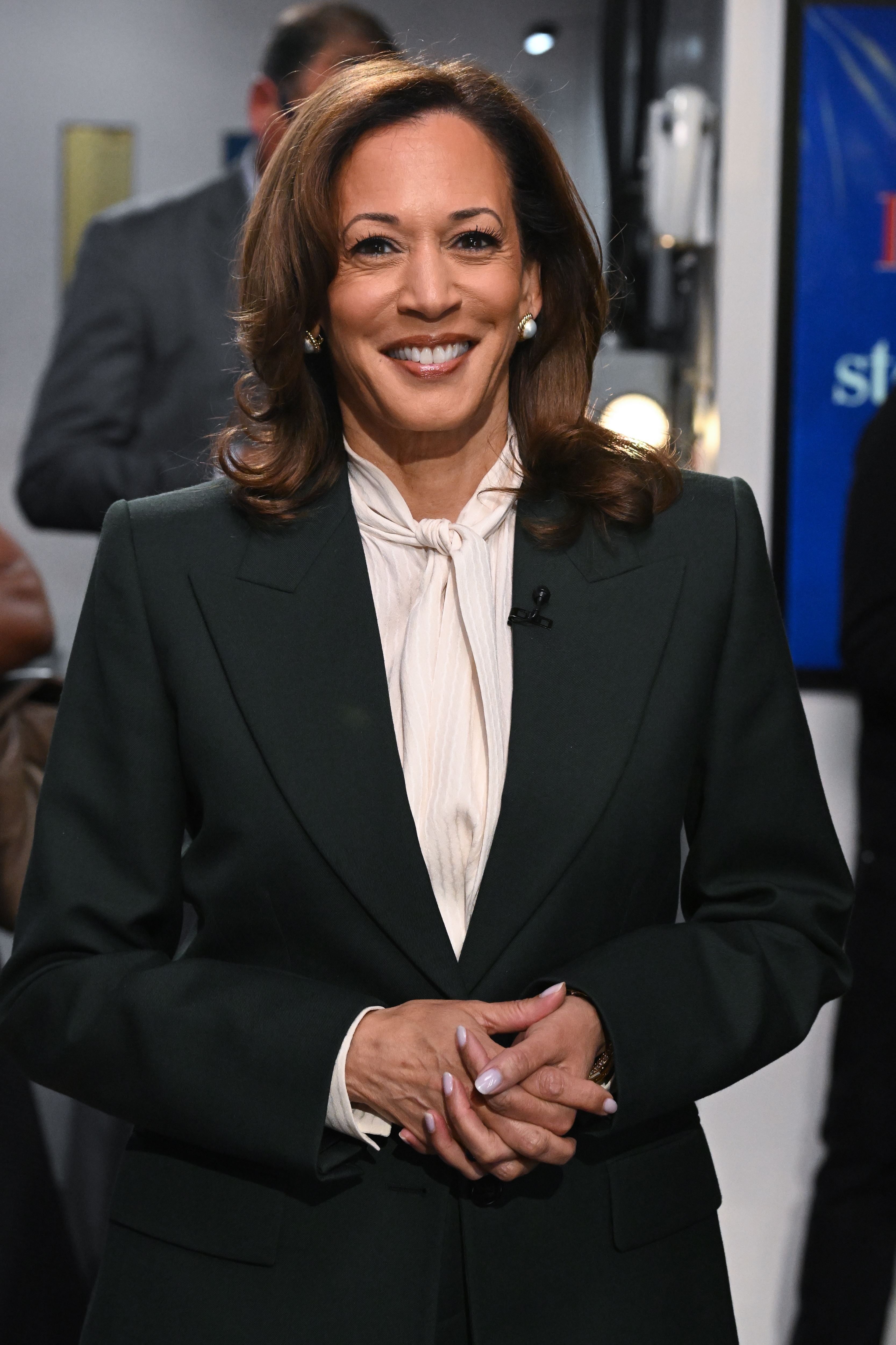 Kamala Harris in a professional suit poses smiling with hands clasped. She stands in a well-lit interior setting