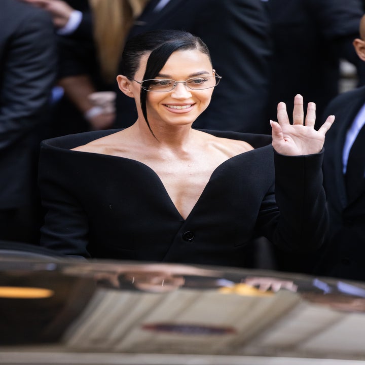 A person in an elegant off-the-shoulder suit waves at an event, wears glasses and sports a stylish hairstyle